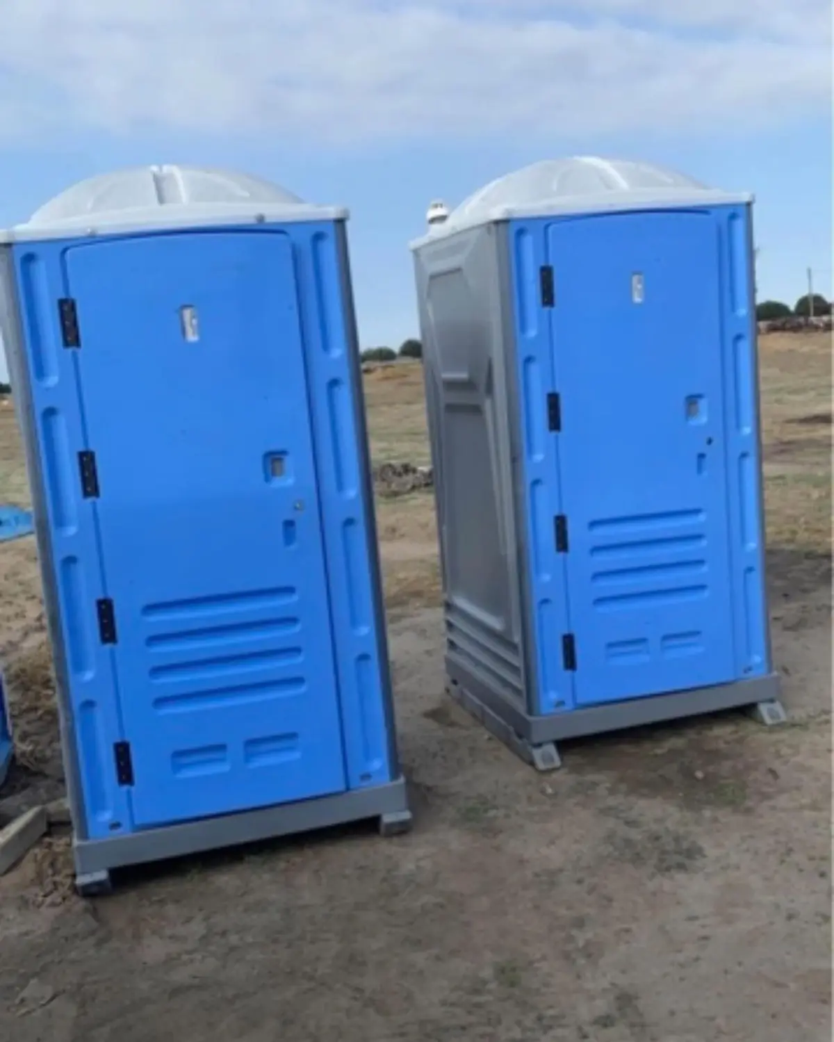 Two blue standard porta potties set side by side on a dirt job site